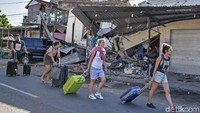 Para turis berjalan membawa kopernya di depan rumah warga yang rusak akibat gempa di Lombok, NTB. (Foto: Antara)