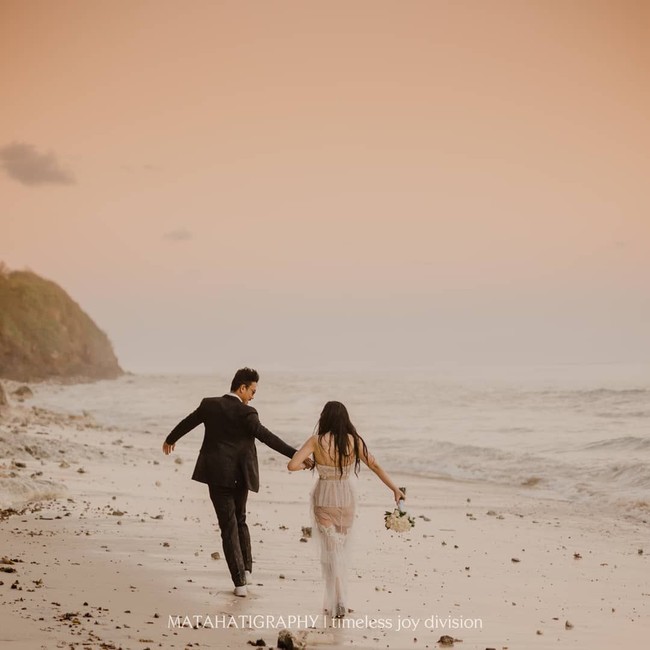 Mesranya Denny dan Dita bergandengan tangan di pantai. Dita yang dibalut gaun putih dan membawa buket bunga, seolah terlihat seperti pengantin yang tengah berbahagia. Foto: Instagram @matahatigraphy