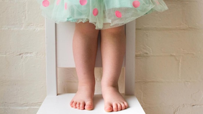 Toddler girl in green and pink polka dot skirt standing on white chair against white brick wall (cropped)