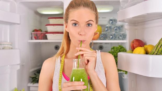 Young beautiful woman standing in the refrigerator door in fitness clothes and drinking green juice while looking at the camera.