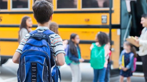 Rear view of elementary age boy waiting to get on school bus. His classmates are loading the bus in the background.