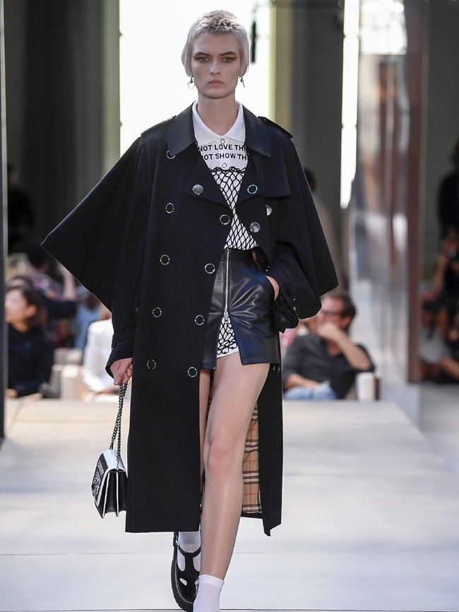 Referensi punk juga muncul pada look yang terdiri dari blus putih yang dibungkus terusan berbahan jaring serta rok kulit berkantong, lalu trench coat hitam sebagai luaran.  Trench coat sebagai produk signature Burberry juga mendapat sentuhan baru dengan siluet lengan yang lebih lebar. (Foto: Dok. Vogue)