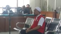 Dia terlihat menunduk dan langsung duduk di kursi terdakwa. Dia terlihat mengenakan baju tahanan berwarna orange terang dan kopiah berwarna putih. 