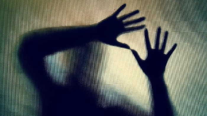 Colour backlit image of the silhouette of a woman with her hands pressed against a glass window. The silhouette is distorted, and the arms elongated, giving an alien-like quality. The image is sinister and foreboding, with an element of horror. It is as if the woman is trying to escape from behind the glass. Horizontal image with copy space.