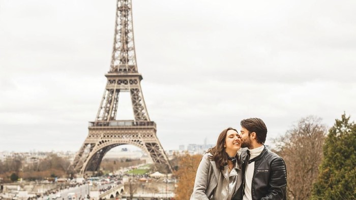 Young couple sitting on surrounding wall with a view on Eiffel tower, the symbol of France. The man kisses his girlfriend on the cheek.