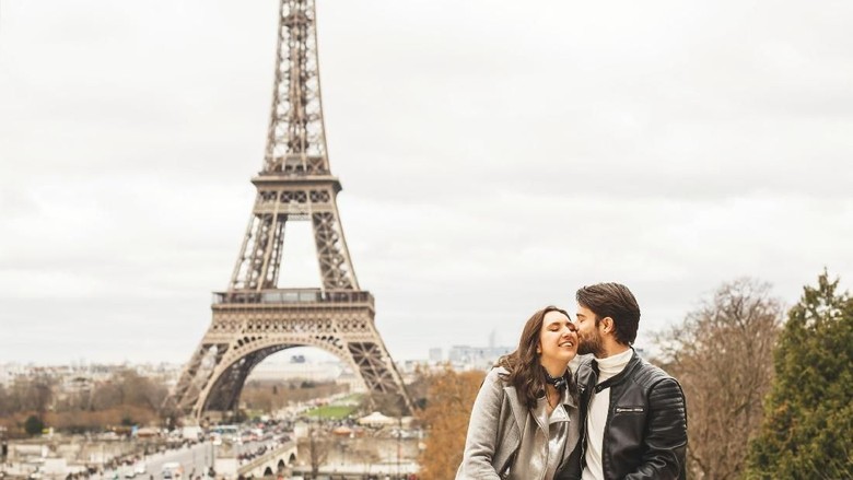 ilustrasi pasangan bulan madu ke paris Young couple sitting on surrounding wall with a view on Eiffel tower, the symbol of France. The man kisses his girlfriend on the cheek.