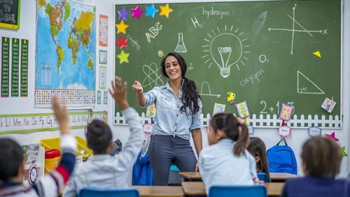An Ethnic teacher is leading a class of elementary school children. There are various posters on the wall, and drawings on the chalkboard. Students are putting up their hands to answer a question.