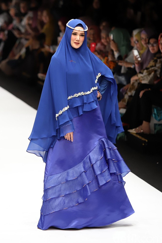 Mulan berlenggak-lenggok di catwalk Jakarta Fashion Week 2019 dalam balutan dress berdetail ruffle berwarna biru. Dia memakai khimar yang sewarna dengan bajunya. Wanita yang kini menjadi istri Ahmad Dhani itu melengkapi gayanya dengan memakai kalung mutiara sebagai hiasan kepala. Foto: Ismail/detikFoto