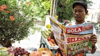 Seorang pedagang buah menunggu pembeli sambil membaca Tabloid Bola edisi terakhir.