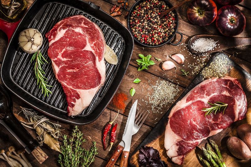 Top view of an iron grill with two fresh beef steak fillets on wooden kitchen table filled with cooking and seasoning ingredients. DSRL studio photo taken with Canon EOS 5D Mk II and Canon EF 100mm f/2.8L Macro IS USM