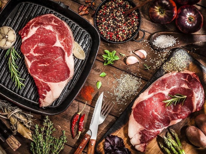 Top view of an iron grill with two fresh beef steak fillets on wooden kitchen table filled with cooking and seasoning ingredients. DSRL studio photo taken with Canon EOS 5D Mk II and Canon EF 100mm f/2.8L Macro IS USM