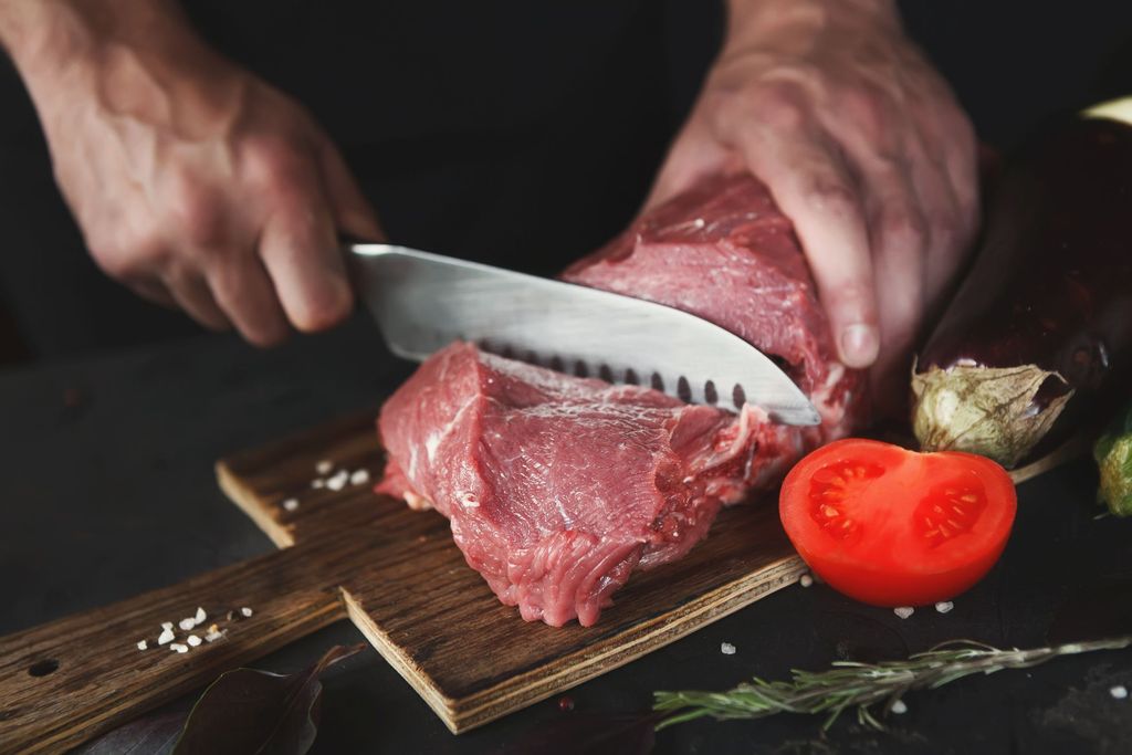 Man slicing filet mignon on wooden board at restaurant kitchen. Chef preparing fresh meat for cooking. Modern cuisine backgroung with herbs and tomato, copy space