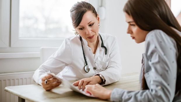 An Asian girl is indoors in a hospital room. She is wearing casual clothing. An Asian female doctor wearing medical clothing is checking her heartbeat with a stethoscope.