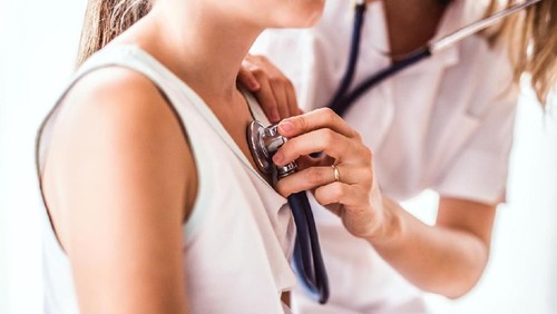 An Asian girl is indoors in a hospital room. She is wearing casual clothing. An Asian female doctor wearing medical clothing is checking her heartbeat with a stethoscope.