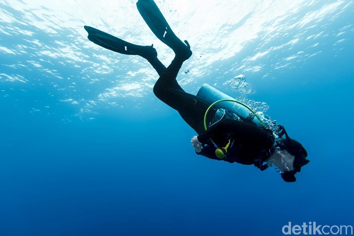 A DSLR underwater photo of a woman scuba diving in Tiputa Pass, Rangiroa, French Polynesia. She is tilted down swimming to the bottom. Above her is the water surface brightly lit by sunshine. The water is superbly clear turquoise blue.