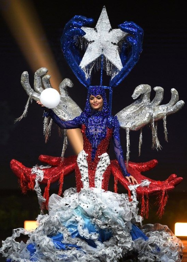 Kostum nasional spektakuler ditampilkan Miss Puerto Rico 2018 Kiara Liz Ortega. Wanita berusia 25 tahun itu memakai bertajuk Puerto Rico Renace. Kiara tampil dalam balutan gaun berwarna biru, merah dan putih dan bagian punggungnya tampak dihiasi empat tangan berwarna merah dan perak. Sementara bagian kepalanya ada dua tangan memegang bintang. Kostum nasionalnya ini merupakan gambaran dari bencana alam Badai Maria yang merenggut banyak korban jiwa di Puerto Rico. Kedua tangan di bagian atas kepalanya merupakan gambaran gotong royong yang dilakukan masyarakat di negaranya untuk bangkit pascabencana badai. Foto: Lillian SUWANRUMPHA / AFP
