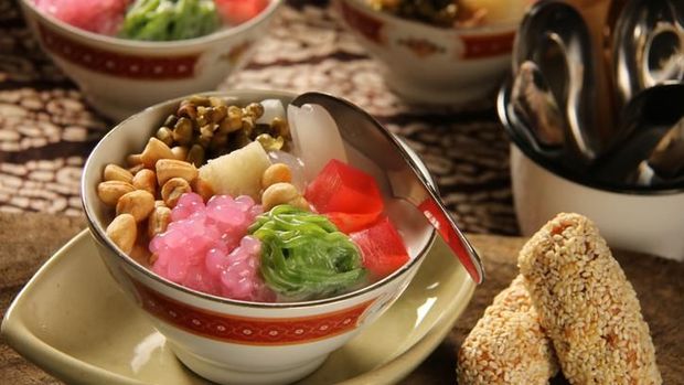 Wedang Angsle, the traditional Javanese warm dessert from Malang, East Java. Tapioca pearls, rice noodle cake, agar jelly cubes, palm fruit, mung beans, peanuts and bread cubes in warm infusion of ginger and coconut milk. The dish is plated in a ceramic deep bowl with a metal soup spoon inserted in the bowl. The dish is served with sticky rice and sesame cakes, a traditional local snack popular in the area. A batik cloth is used as a table cloth.