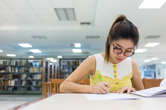 Female student with long hair sitting in the library while writing on the book with laptop at desk