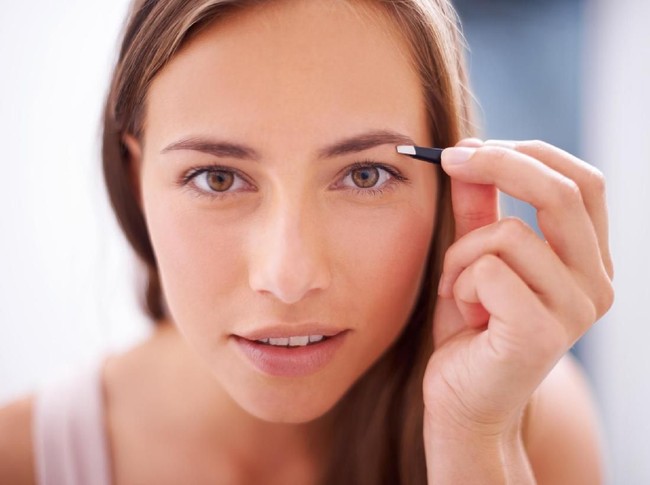 Closeup of attractive mid 20s blond woman putting on some makeup in front of large bathroom mirror. Shot from behind, focus is on her reflection. Softly toned and desaturated.