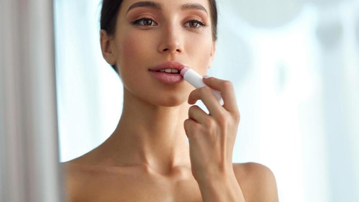 Closeup of attractive mid 20s blond woman putting on some makeup in front of large bathroom mirror. Shot from behind, focus is on her reflection. Softly toned and desaturated.