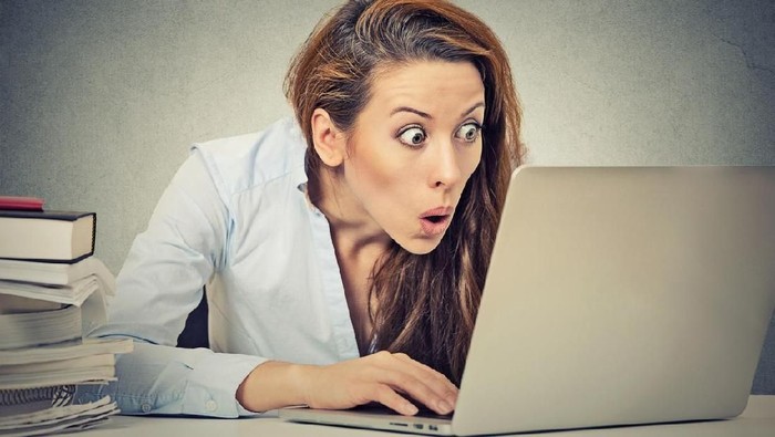 Portrait young shocked business woman sitting in front of laptop computer looking at screen isolated on grey wall background. Funny face expression emotion feelings problem perception reaction