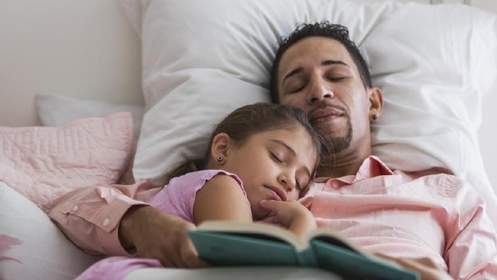 african american father holding book and palming sleeping daughter at home