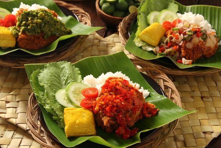 Ayam Geprek, the popular fusion street food dish of southern fried chicken served with traditional spicy condiments from regional cuisines in Indonesia. Here each of the chicken piece is topped with a popular condiment, such as Sambal Lado (lightly red chili paste) and Sambal Lado Mudo (green chili paste), both from West Sumatra, and Sambal Matah (chili pepper, lemon grass and shallot salsa) from Bali. The chicken and condiment are served with steamed rice, yellow tofu, and fresh vegetables. The dish is plated on a ceramic plate lined with banana leaf; which then placed on a woven stick plate.
