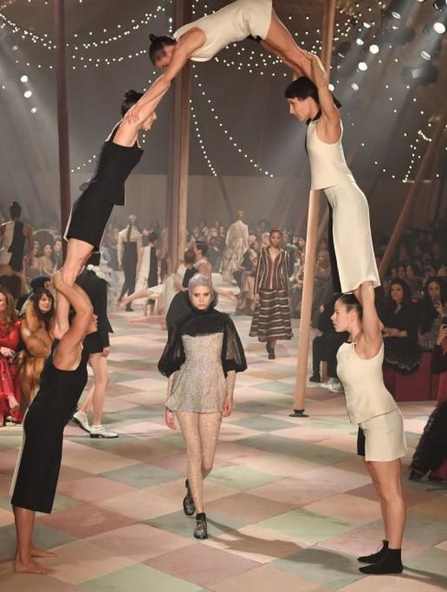 Para pemain akrobat membentuk formasi piramida yang menyimbolkan kekuatan seorang perempuan. Model dalam balutan gaun mini transparan berjalan di bawahnya. (Foto: Pascal Le Segretain/Getty Images For Christian Dior)