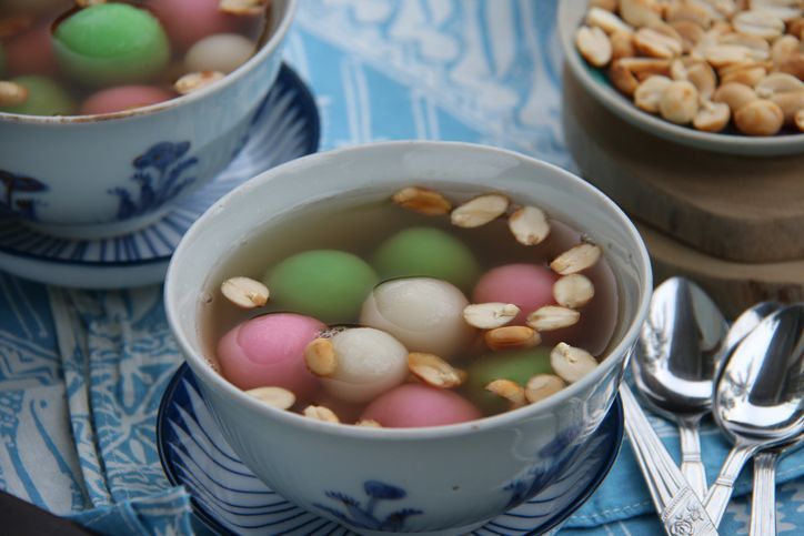 A Chinese Peranakan dessert of glutinous rice balls in warm ginger soup. Served in vintage ceramic bowls with extra peanuts, also in vintage saucer. A few teaspoons are placed on a folded batik napkin. The meal is arranged on a rustic wooden table.