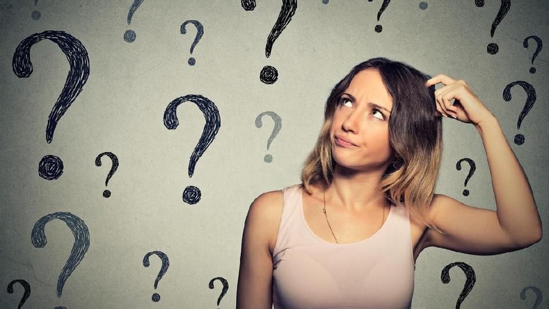 Thinking young woman looking up at many question marks isolated on gray wall background