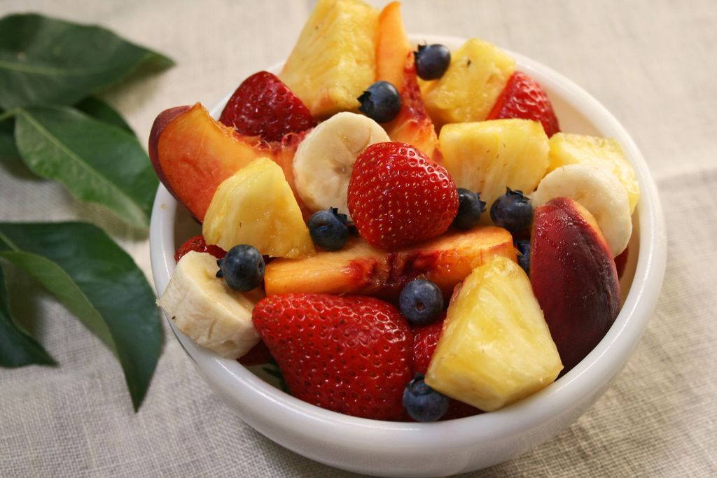 A selection of different Summer fruits, in a variety of bowls on a painted blue wood planked farmhouse kitchen table, against a light blue background.