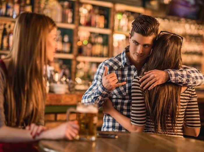 Unfaithful young man in a cafe. He is embracing his girlfriend and making call gesture to another woman behind her back.