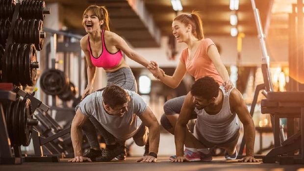 Cheerful men doing push-ups in a health club while women are sitting on their back's and laughing. Focus is on men.
