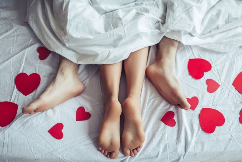 Cropped image of young couple is lying on bed. Close up of male and female feet. Loving couple is lying on bed under blanket covered by small red paper hearts. Saint Valentines Day.