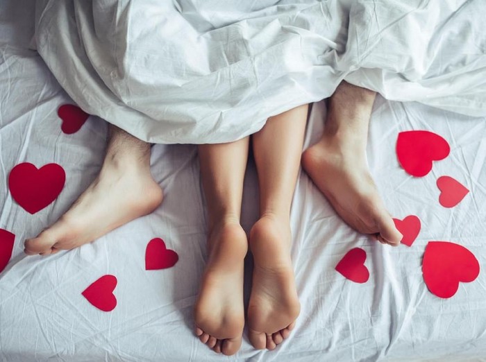 Cropped image of young couple is lying on bed. Close up of male and female feet. Loving couple is lying on bed under blanket covered by small red paper hearts. Saint Valentines Day.