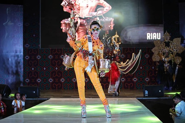 Memilih blus dan celana panjang serba oranye, Lycie Joanna asal Kepulauan Riau menyempurnakan gayanya dengan penutup mata bak superhero. Busananya juga dihiasi peralatan musik berwarna emas yang mempertegas karakter budaya Melayu di Riau.  Foto: Instagram/@officialputeriindonesia