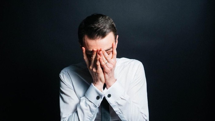 ilustrasi pria sedih Color portrait of a young man in a shirt and black tie, upset, hands on his face, against plain studio background.