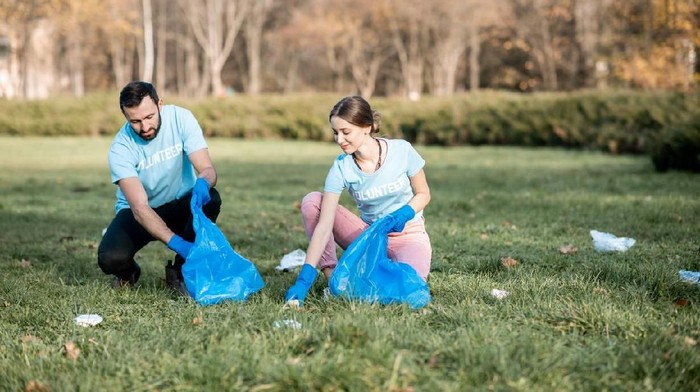 Happy man and woman volunteers dressed in blue t-shirts cleaning public garden from plastic rubbish into the bags