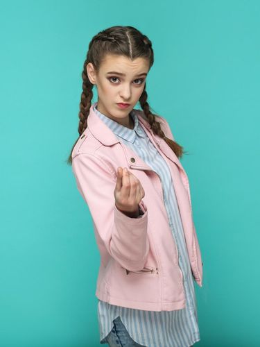 money gesture. portrait of beautiful cute girl standing with makeup and brown pigtail hairstyle in striped light blue shirt pink jacket. indoor, studio shot isolated on blue or green background.