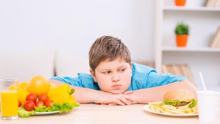Hard choice. Chubby kid is sitting between two plates with different kind of food.