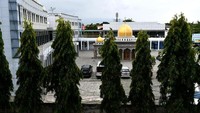 Beberapa pohon tumbuh di depan gedung. Para pegawai beraktivitas di sana. Pada gedung bagian depan, dindingnya terlihat dikelilingi kaca biru.