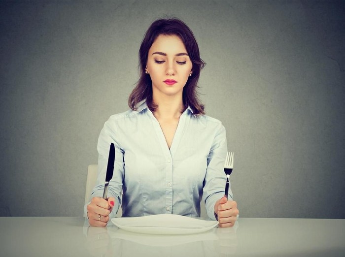 Serious woman with fork and knife sitting at table with empty plate isolated on gray wall background