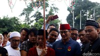 Sandi nampak mengenakan kopiah berwarna merah dan mengenakan pakaian berwarna biru saat tiba di GOR Ciracas, Jakart Timur.