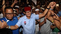 Sandi nampak mengenakan kopiah berwarna merah dan baju berwarna biru muda saat tiba di Gelanggang Remaja Jakarta Utara, Tanjung Priok, untuk mengisi kampanye akbar. Ia pun tersenyum menyapa para simpatisan yang hadir.