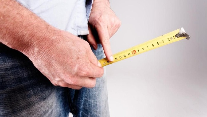 An unrecognizable mans hands hold a tape measure to the crotch of his jeans, checking size or growth with his forefinger.