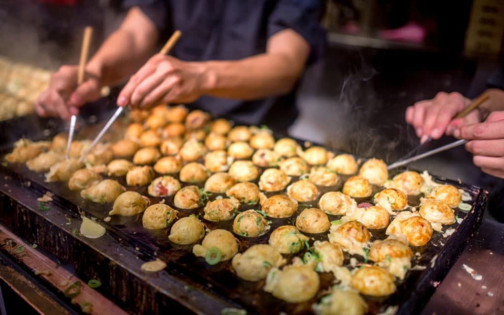 young asian woman holding box of japanese local street food looking around. lady traveler enjoy eating takoyaki on the road outdoor. delicious octopus balls in osaka japan.