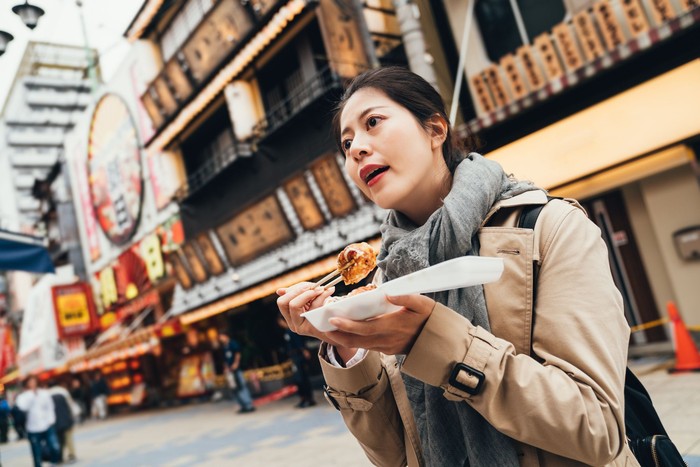 Larang Warga Makan di Jalanan young asian woman holding box of japanese local street food looking around. lady traveler enjoy eating takoyaki on the road outdoor. delicious octopus balls in osaka japan.