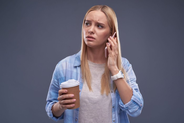 Waist up portrait of charming blond girl holding cup of coffee and having phone conversation while looking away with concerned expression. Isolated on blue-gray background