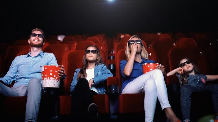 Closeup front view of young family with two daughters watching a 3D movie at movie thater. They are sitting on first row, eating pop corn and wearing 3D glasses.