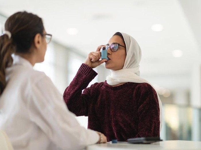 A Muslim woman wearing a hijab sits at a table while at a medical consultation. She has asthma and is learning to use an inhaler. A female doctor is watching and instructing the patient on how to use a puffer. The doctors back is to the camera.